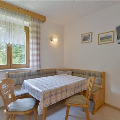 A cozy dining area with a table and a bench. The walls are light and there are windows with curtains as well as some pictures.