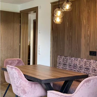 Ein eleganter Essbereich mit einem Holztisch und rosa Stühlen. Die Wände sind in einem warmen Holzton gehalten und die Beleuchtung schafft eine gemütliche Atmosphäre.