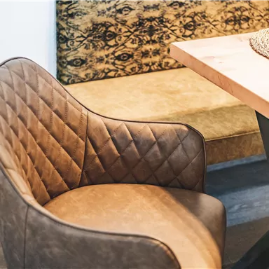 Ein eleganter Stuhl mit einer gepolsterten, hellbraunen Oberfläche steht neben einem Tisch. Im Hintergrund ist eine gemütliche Bank zu erkennen.