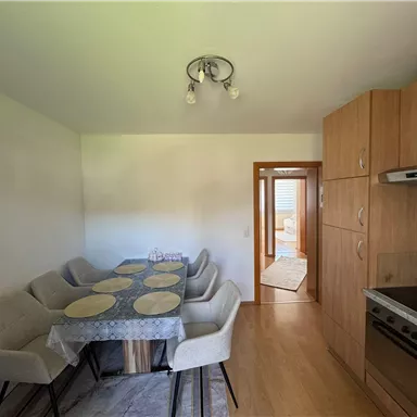 A modern kitchen with a large dining table and beige chairs. In the background, there is a door that leads to another room.