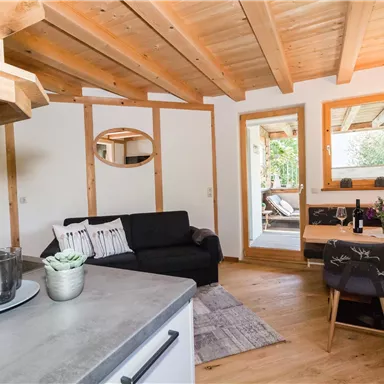 Eine moderne, gemütliche Wohnküche mit Holzboden und viel natürlichem Licht. Es gibt ein Sofa, einen Esstisch und Zugang zu einer Terrasse.