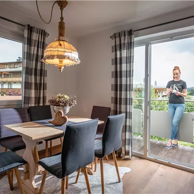 A bright dining room with an elegant table and black chairs. A person is standing on the balcony and looking outside.