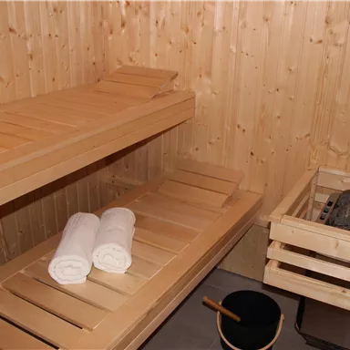 A cozy wooden sauna with two benches and clean towels. Next to the sauna bench are a sauna stove and a bucket.