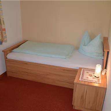 A simple bedroom with a single bed and a bedside table. The walls are painted light, and there is a window with curtains.