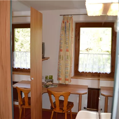 A cozy room with two tables and chairs. Next to a window with curtains, some plants and a television are visible.