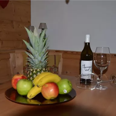 Ein Tisch mit einer Obstschale, die Ananas, Äpfel, Birnen und Bananen zeigt. Daneben steht eine Flasche Weißwein und Gläser.