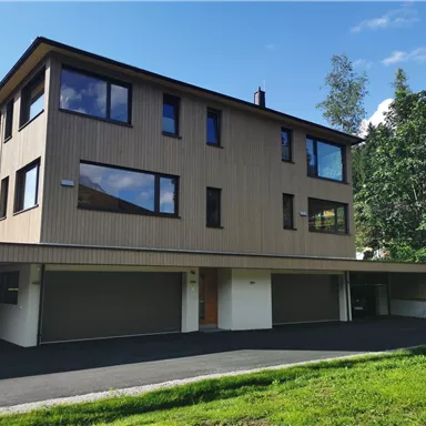 Ein modernes Haus mit Holzfassade und großen Fenstern. Im Vordergrund sind Garagen und eine grüne Rasenfläche zu sehen.