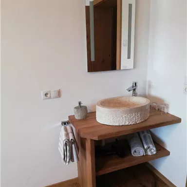 Ein modernes Badezimmer mit einem Holzwaschtisch und einer runden Waschschüssel. Im Hintergrund ist ein Spiegel sowie Handtücher und Gläser auf dem Regal zu sehen.
