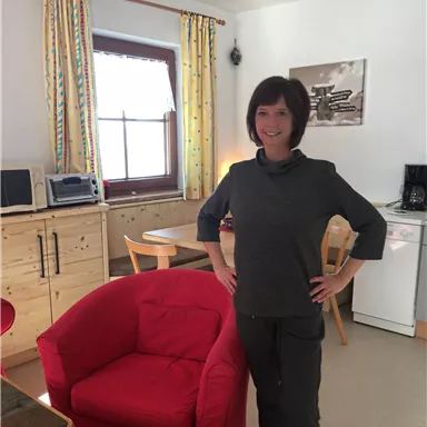 A woman stands in a cozy kitchen with modern furniture. In the background, a red armchair and a table can be seen.