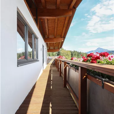 A bright balcony with a wooden floor and flower boxes. In the background, mountains and a blue sky can be seen.