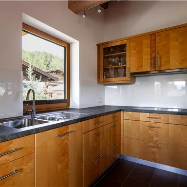 A modern kitchen with wooden cabinets and a large window view. The countertop is made of black material and offers plenty of space for cooking.