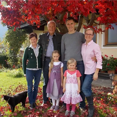 Eine fröhliche Familie steht zusammen im Garten unter einem roten Baum. Im Hintergrund sind Blumen und ein Haus sichtbar.