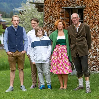 A group of five people is standing outdoors in front of a stack of wood. They are dressed in traditional clothing and are smiling kindly at the camera.