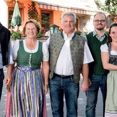 A group of five people in traditional clothing stands together. In the background, a few green plants and a building are visible.