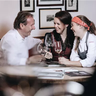 Two women and a man are sitting at a table, laughing and toasting with wine glasses. In the background, there are pictures on the wall.