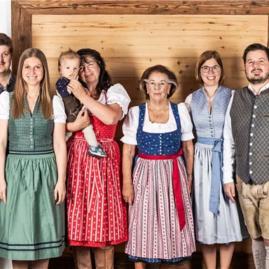 A group of six people in traditional Bavarian attire stands in front of a wooden wall. They are smiling and appear friendly.