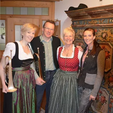 Eine Gruppe von vier Personen in traditioneller Tracht steht in einem gemütlichen Raum. Im Vordergrund sind rote Rosen und ein dekorativer Schrank zu sehen.