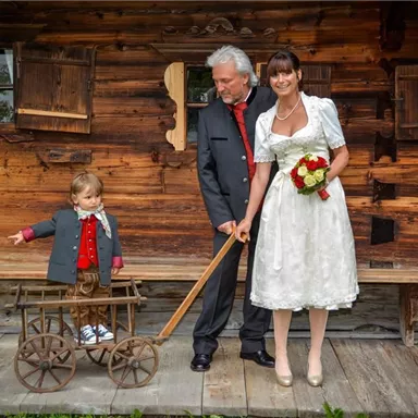 Ein Brautpaar steht vor einem Holzhaus. Der Mann trägt einen schwarzen Anzug, die Frau ein weißes Kleid, während ein kleiner Junge mit einem Wagen spielt.