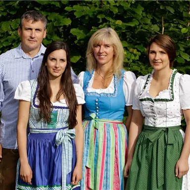 Eine Gruppe von vier Personen, die traditionelle bayerische Trachten tragen. Sie stehen lächelnd vor einer grünen Kulisse.
