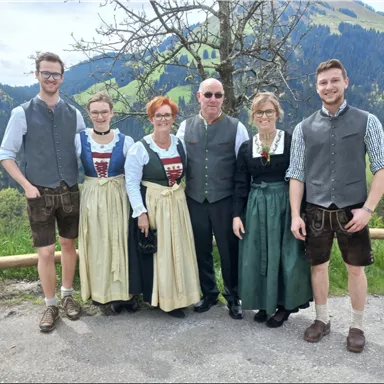 A group of six people in traditional costumes stands in front of a picturesque mountain landscape. They are smiling and posing against a backdrop of green hills and trees.