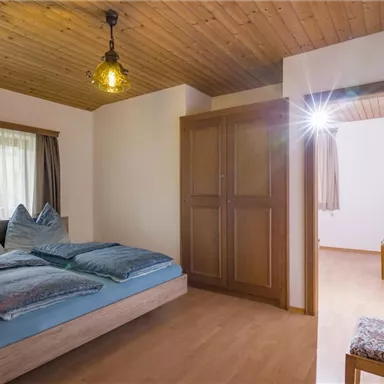Ein gemütliches Schlafzimmer mit einem Doppelbett, blauen Kissen und einer warmen Holzdecke. Das Zimmer ist hell und hat einen Kleiderschrank sowie einen Ausblick in einen angrenzenden Raum.