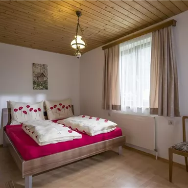 Ein gemütliches Schlafzimmer mit einem Doppelbett und roten Kissen. Das Zimmer hat Holzdecken und ein Fenster mit Vorhängen.