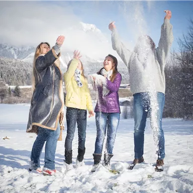 Eine Gruppe von vier Freunden steht in einem verschneiten Landschaft und wirft Schnee in die Luft. Sie lachen und genießen die winterliche Stimmung.