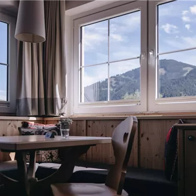 A cozy room with a wooden table and chairs. Through the windows, you can see the mountains and the blue sky.