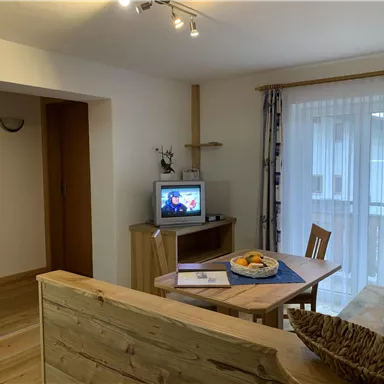 A cozy living space with a table, chairs, and a television. There are large windows that let in a lot of light.