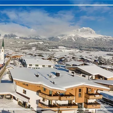 A picturesque winter landscape with snow-covered roofs and mountains in the background. A church tower rises into the clear blue sky.