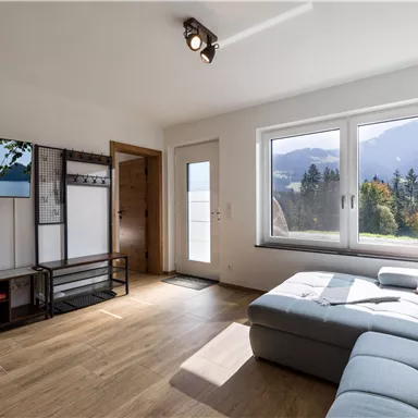 A bright living room with large windows and a view of the mountains. The decor is modern and inviting, with a cozy sofa and a TV corner.