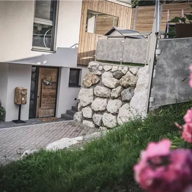 Ein modernes Haus mit einer Steinmauer und einem schönen Eingang. Im Vordergrund blühen pinke Blumen.