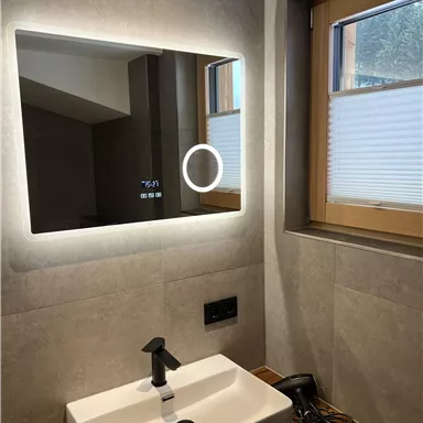A modern bathroom with a lit mirror and a white sink. Natural light is coming in through the window.