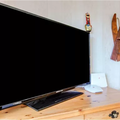 Ein schwarzer Fernseher steht auf einem Holztisch in einem gemütlichen Raum. Im Hintergrund sind dekorative Skier und eine kariierte Wand zu sehen.