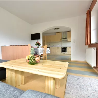 A cozy living room with a modern kitchen in the background. A wooden table with fruit is at the center of the room.