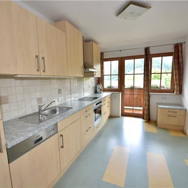 A modern kitchen with light wooden furniture and large windows. The room has a practical layout and ample space for meal preparation.