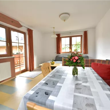 A cozy living space with a dining table and a beautiful flower vase. Large windows let in plenty of light and offer a view outside.