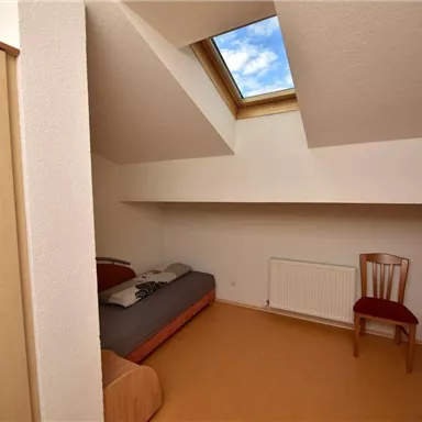A simple room with two single beds and a chair. A window in the roof provides daylight and a view of the sky.