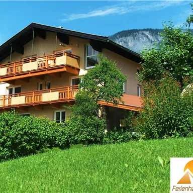 A beautiful holiday home with a balcony, surrounded by green grass and trees. Mountains can be seen in the background.