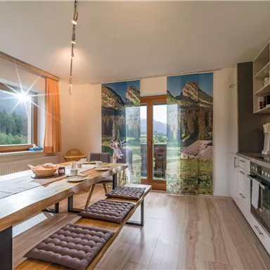 Eine moderne Küche mit einem großen Esstisch und Blick auf die Berge. Helle Farben und große Fenster sorgen für eine einladende Atmosphäre.