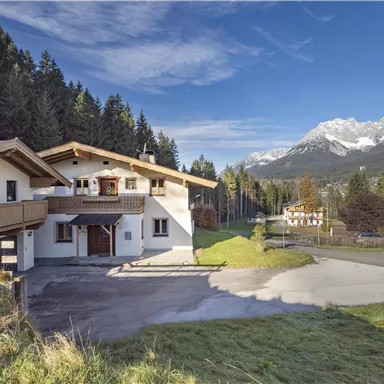 A beautiful house in the mountains, surrounded by trees and with snow-capped peaks in the background. The sky is clear and blue.