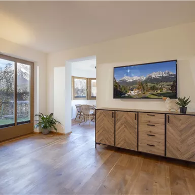 A modern living room with wooden flooring and a television on the wall. Large windows provide a view outside and plenty of natural light.