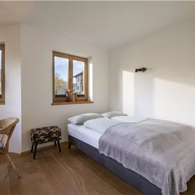 A bright bedroom with two windows and a view of the mountains. There is a bed, a rattan chair, and a small stool.