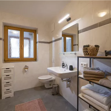 A modern bathroom with bright tiles and a large mirror. Towels are neatly stacked on a shelf, and there is a window with natural light.