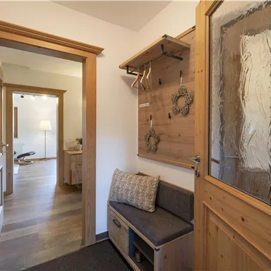 A bright hallway with wooden paneling and a cozy bench. On the wall are coat hooks and a glass door leading to another room.