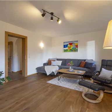 A cozy living room with a gray couch, a wooden coffee table, and a floor lamp. A colorful painting hangs on the wall, and the floor is wooden.