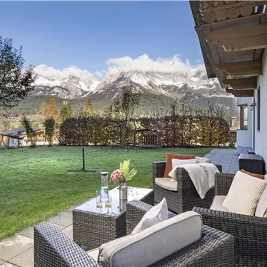 A cozy terrace with rattan furniture and a table in the foreground. Majestic mountains and a clear sky are visible in the background.