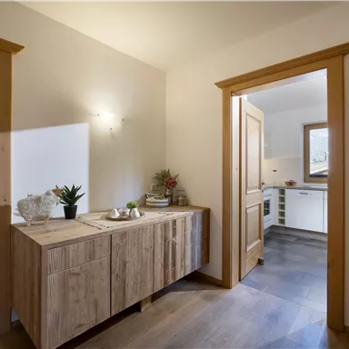 A bright hallway with a modern wooden wardrobe and a plant arrangement. In the background, an open door to the kitchen is visible.