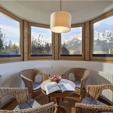 A cozy interior with a table and four rattan chairs. The large windows offer a view of the mountains and nature outside.