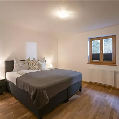 A cozy bedroom with a large bed and bright walls. The window lets in plenty of daylight and the floor is made of wood.
